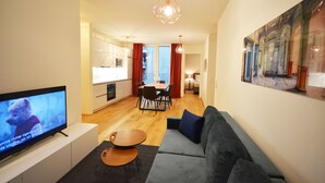 A modern living room with a flat-screen TV, a grey sofa, a wooden dining table with chairs, and a kitchen in the background. - Premium Residence near Parliament (Vienna)