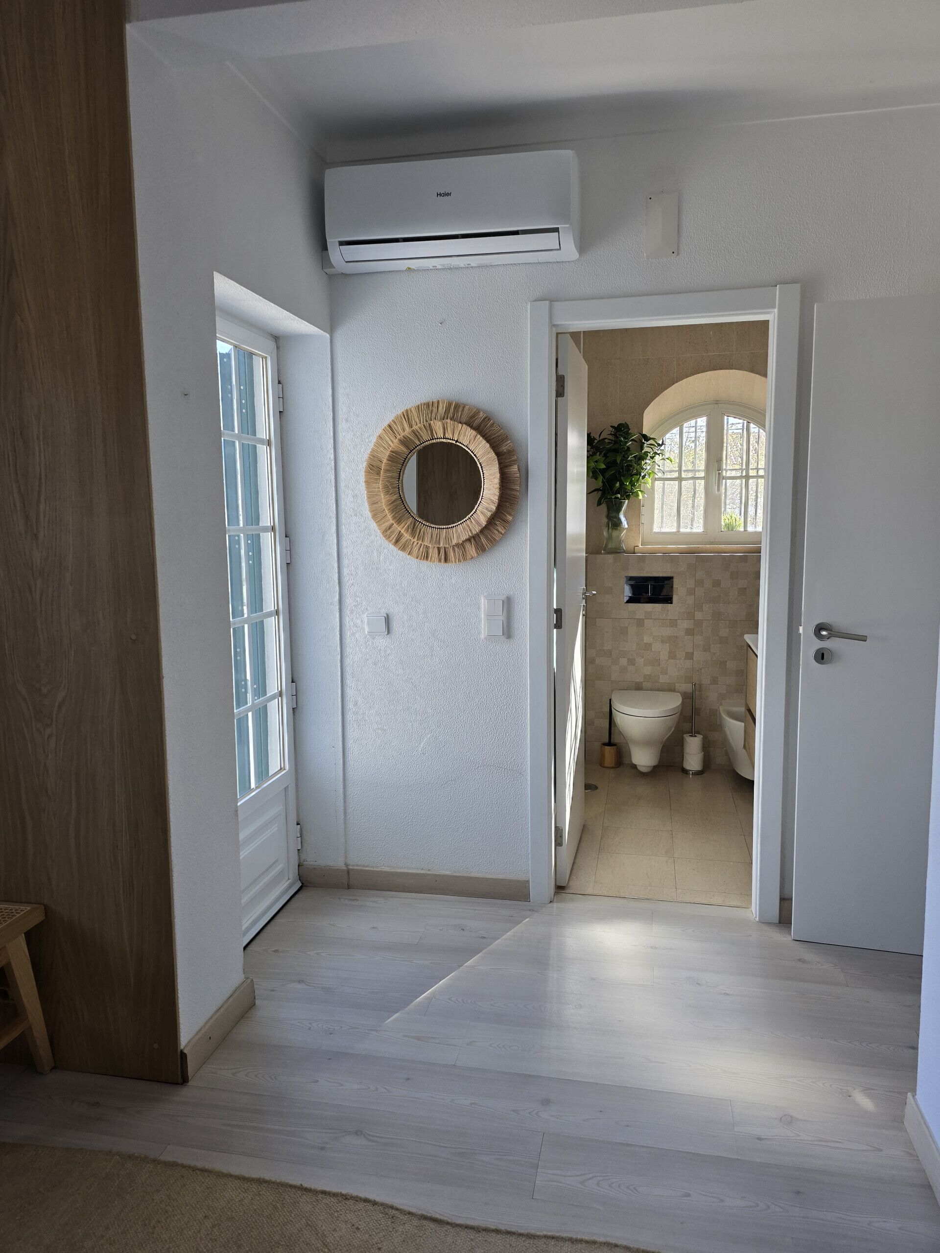Corredor que dá para um banheiro com um espelho redondo, um ar-condicionado e uma porta de madeira.