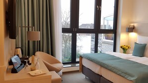 Habitación de hotel con cama, escritorio con computadora y una botella, una silla, ventana con cortinas y vista a edificios.