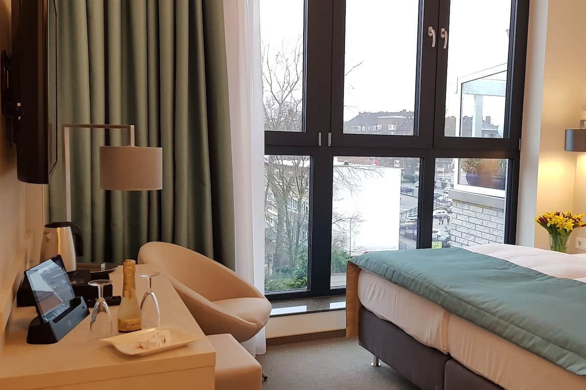 A hotel room with a bed, a desk with a computer and a bottle, a chair, a window with curtains, and a view of buildings outside.