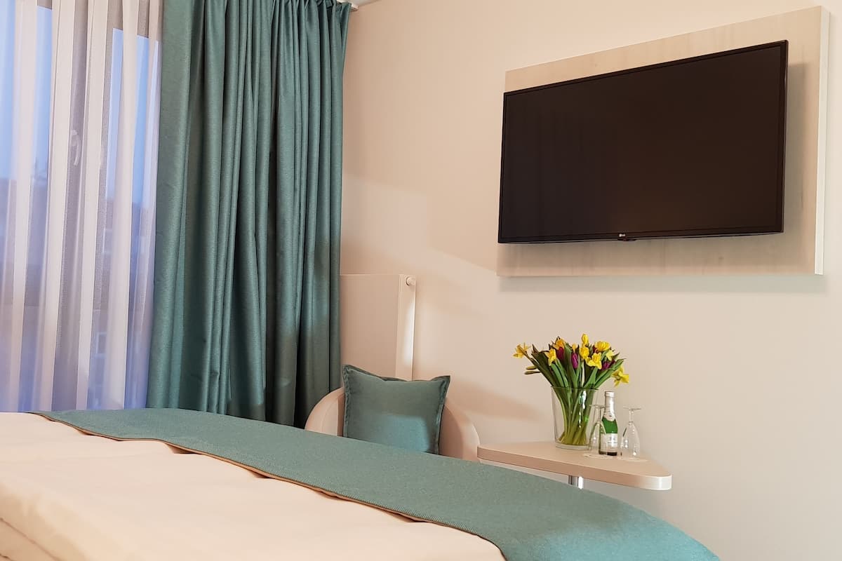 A bedroom with a bed, a TV mounted on the wall, a bedside table with a vase of flowers, and sheer curtains.