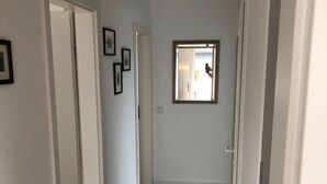 A hallway with wooden flooring, white walls, and a door leading to another room. There are framed pictures on the wall and a small window with a view of the outside.