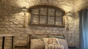 Ein schlichtes Schlafzimmer mit Steinwänden, einem rustikalen Holz-Kopfteil, einem kleinen Nachttisch und einem Fenster mit Holzrahmen.