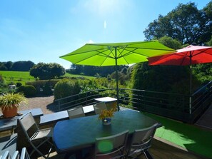 Un patio al aire libre con una mesa y sillas, sombrillas verdes y rojas, y vistas a un paisaje verde.