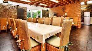 A dining area with a long table and wicker chairs, wooden beams on the ceiling, stone walls, and a view of greenery outside. - La Ferme des Prés (Neuilly-en-Sancerre)