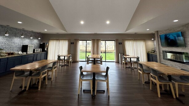 A spacious dining area with multiple wooden tables and chairs, large windows with curtains, and a kitchen area with a brick wall and pendant lighting.
