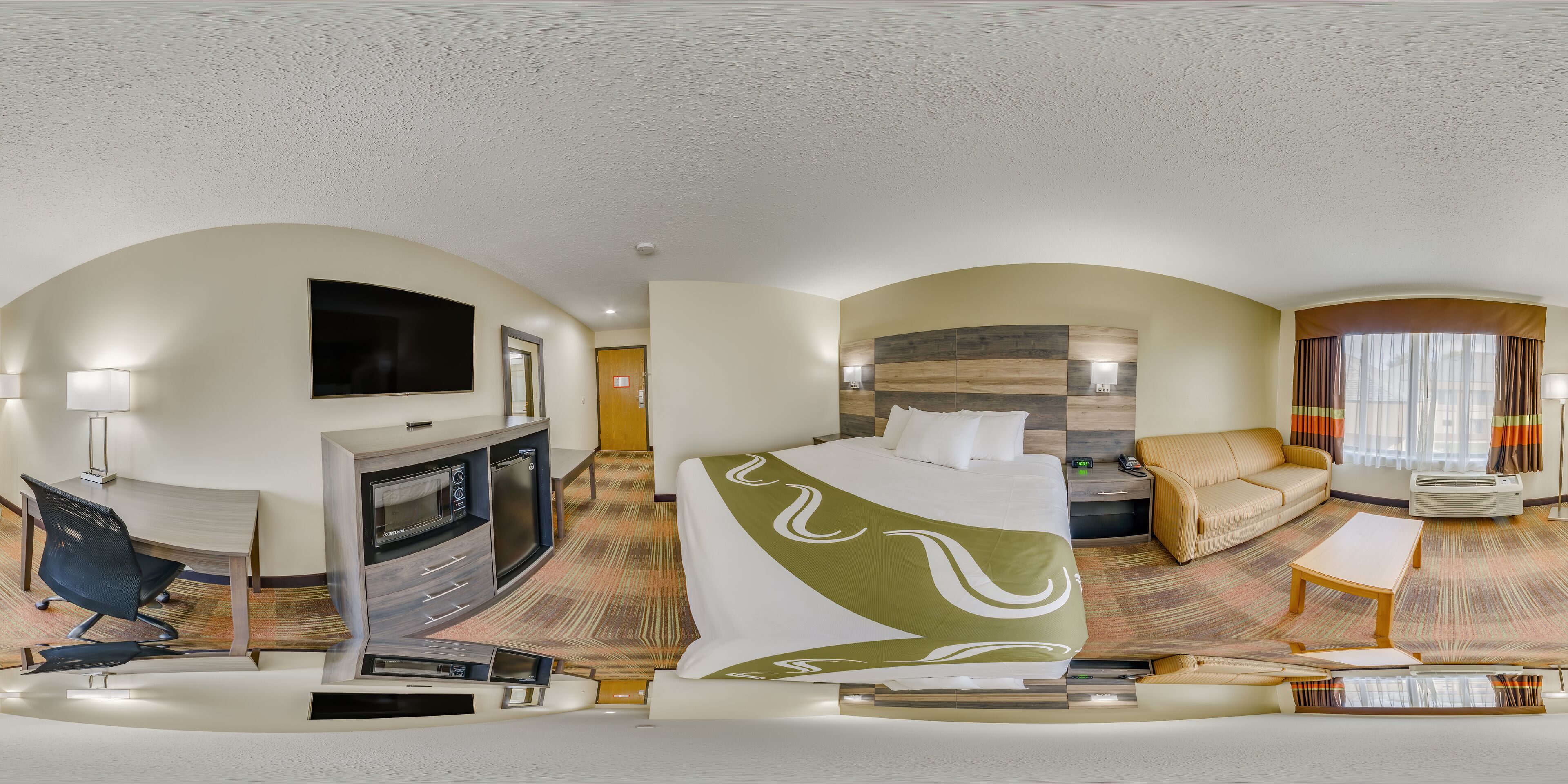 A hotel room with a bed, a sofa, a desk with a TV, and a kitchenette.