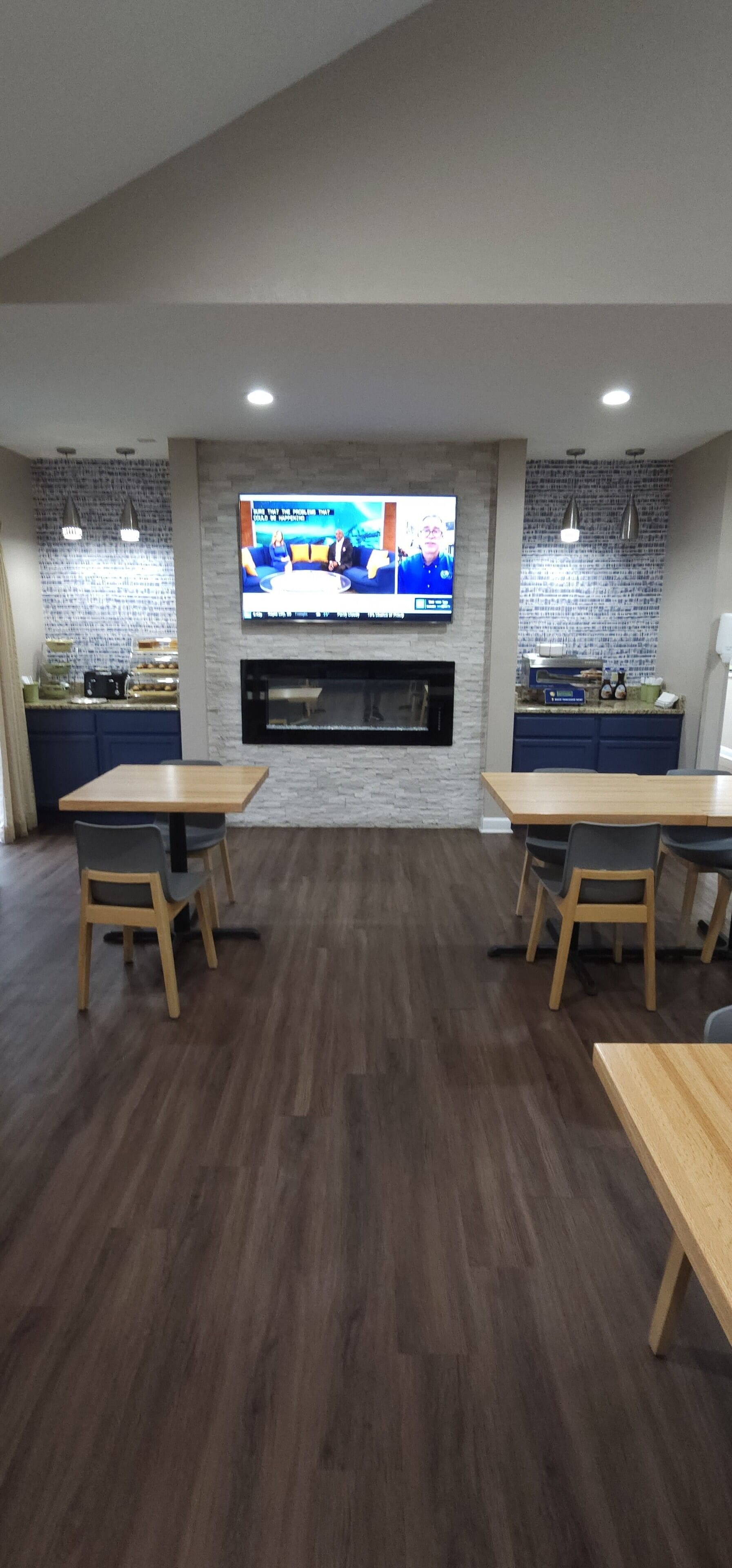 A modern dining area with wooden tables and chairs, a flat-screen TV mounted on the wall, and a kitchen area with a stone backsplash.