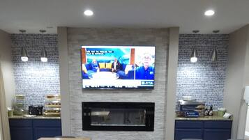 A modern dining area with wooden tables and chairs, a flat-screen TV mounted on the wall, and a kitchen area with a stone backsplash.