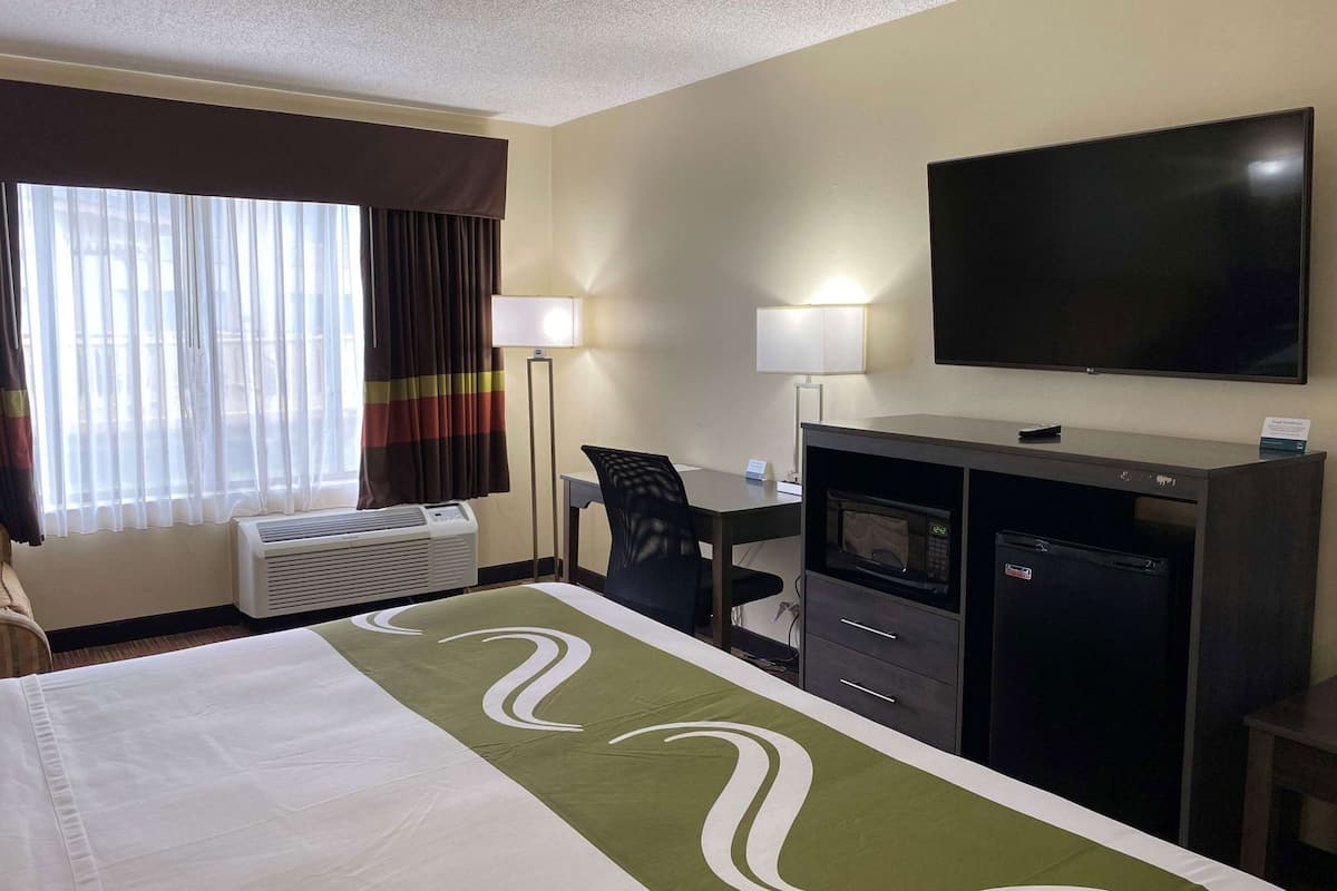 A hotel room with a bed, desk, TV, microwave, and air conditioning unit.