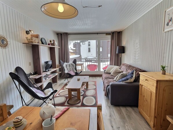 Ein möblierter Wohnraum mit einer Couch, einem Esstisch, einem Fernseher und Blick durch eine Schiebetür.