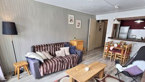 A living room with a sofa, a wooden table, a dining table with chairs, a lamp, and a kitchen area with a refrigerator. - 2-room apartment mountain view - 4 guests (Val-d'Isère)