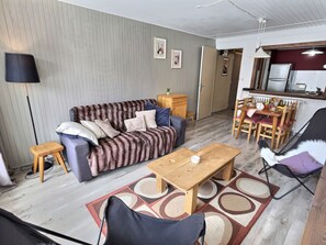 Ein Wohnzimmer mit einer Couch, einem Holztisch, einem Essbereich mit Stühlen, einer Lampe und einer Küchenzeile mit Kühlschrank.