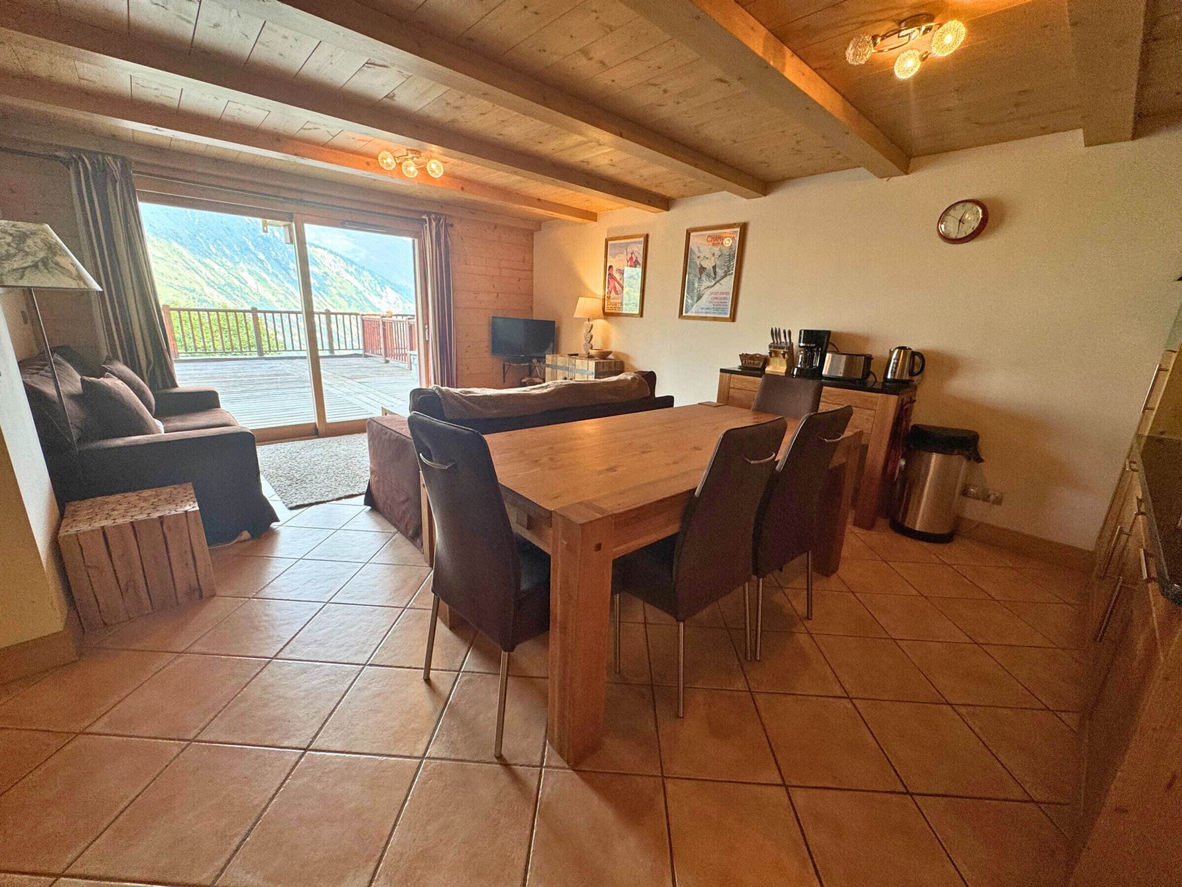 A cozy room with a wooden dining table, chairs, a sofa, and a TV. There's a window with a view of mountains.