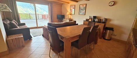 A cozy room with a wooden dining table, chairs, a sofa, and a TV. There's a window with a view of mountains.