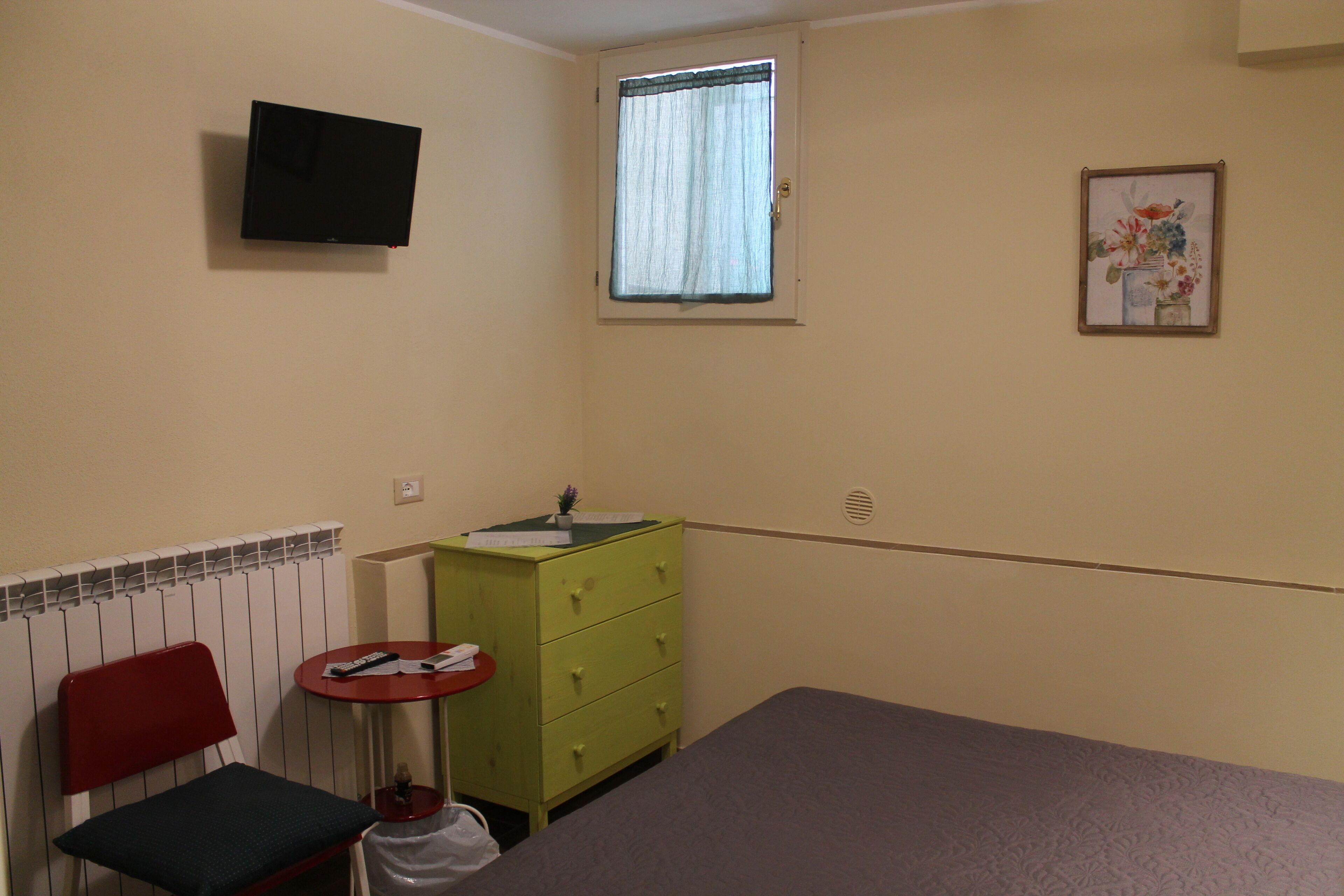 Une petite pièce simple, avec un lit, une chaise, une petite table, une commode verte, une fenêtre avec des rideaux, un téléviseur fixé au mur et un tableau encadré accroché au mur.