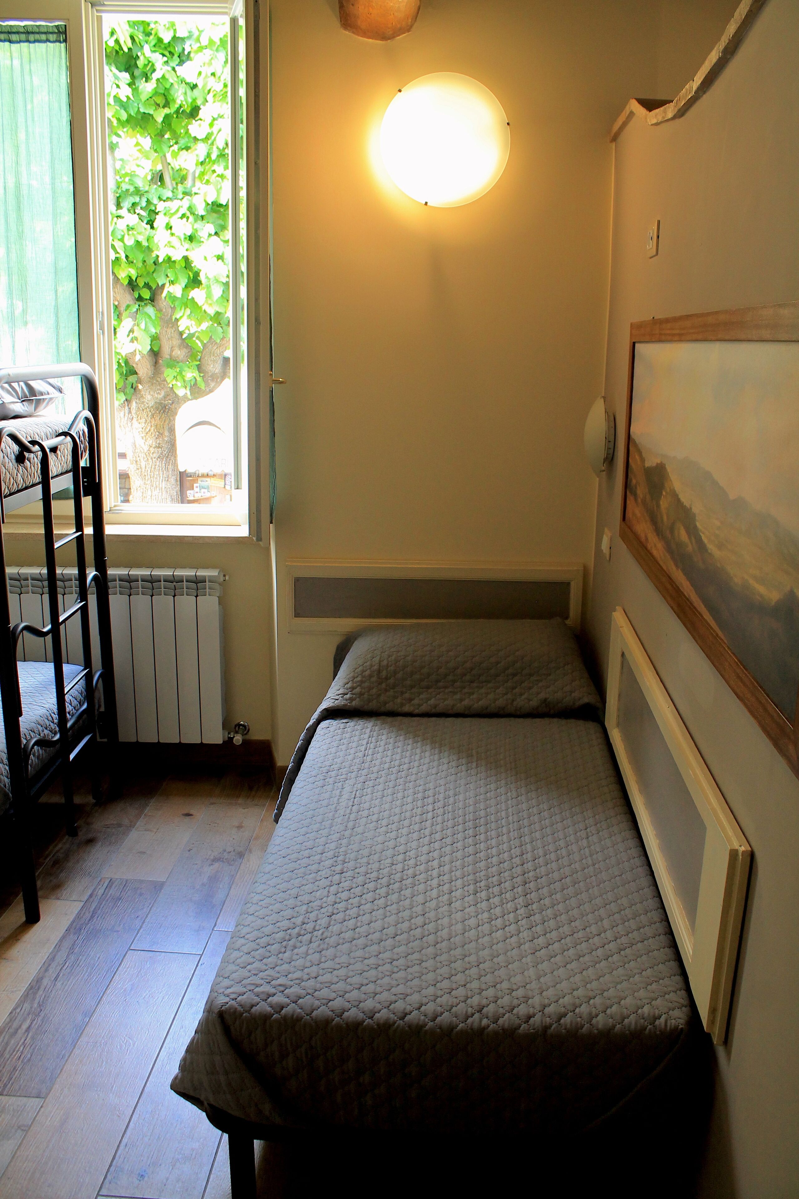 Una habitación pequeña y acogedora con literas, un radiador, una ventana con vistas a un árbol y una lámpara fijada en la pared.
