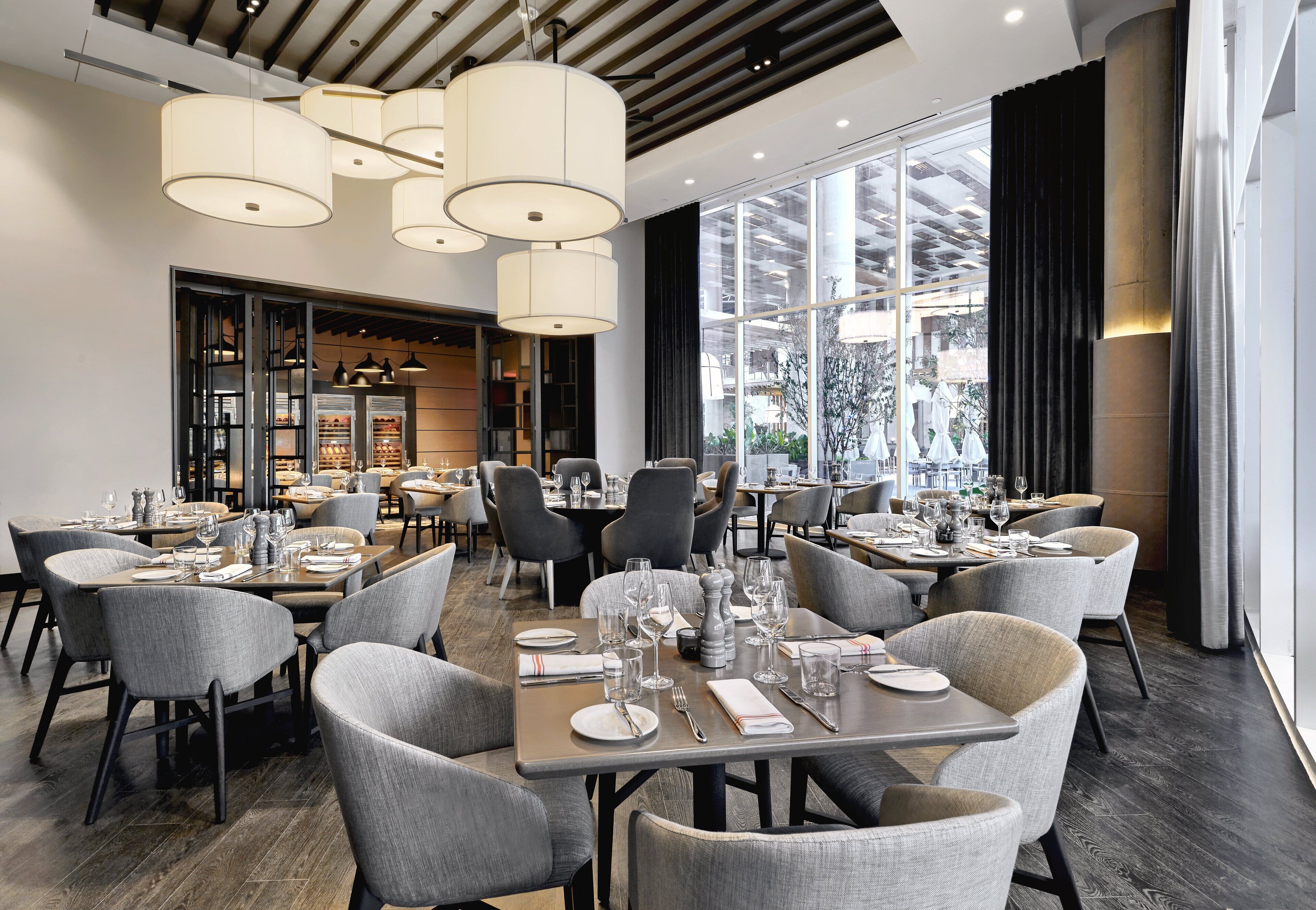 A modern restaurant with multiple dining tables, grey upholstered chairs, and large windows offering a view of the city.