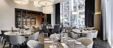 A modern restaurant with multiple dining tables, grey upholstered chairs, and large windows offering a view of the city.