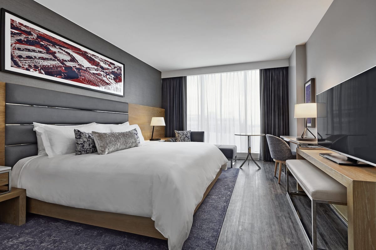 Moderni hotellihuone, jossa on suuri sänky, työp öytä tuolilla, televisio ja suuri ikkuna, jossa on verhot.
