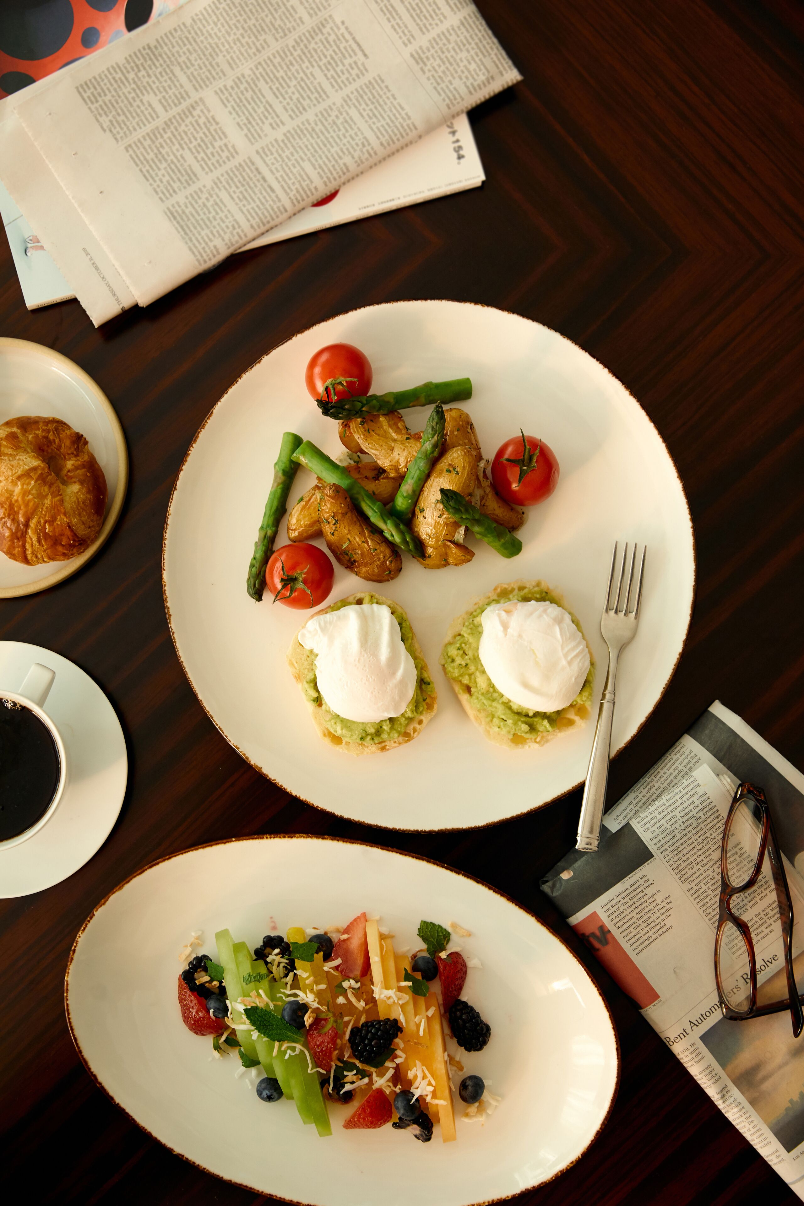 A dining table with two plates of food, a croissant, a cup of coffee, and a newspaper.