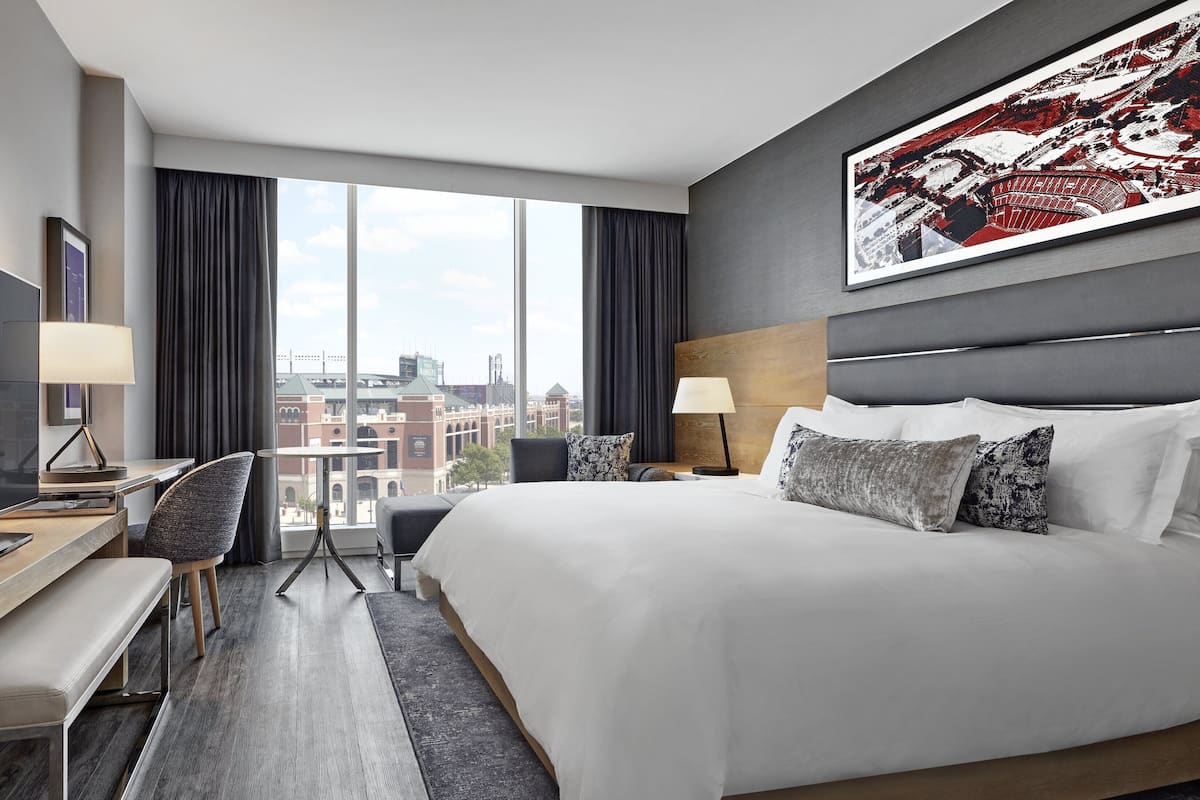 Moderni hotellihuone, jossa on suuri sänky, työpöytä tuolilla, televisio ja suuri ikkuna, josta avautuu näkymä kaupunkiin.