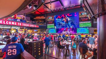A crowded indoor venue with multiple screens displaying live events, a bar area, and a sign welcoming visitors to Texas.