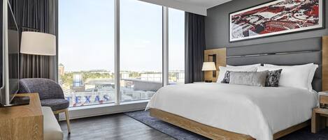 A modern hotel room with a large bed, a desk, a chair, and a large window offering a city view.