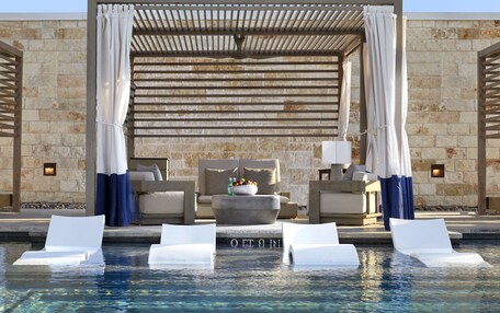 A pool area with white lounge chairs, a stone wall, and a covered seating area with a bowl of fruit and a lamp.