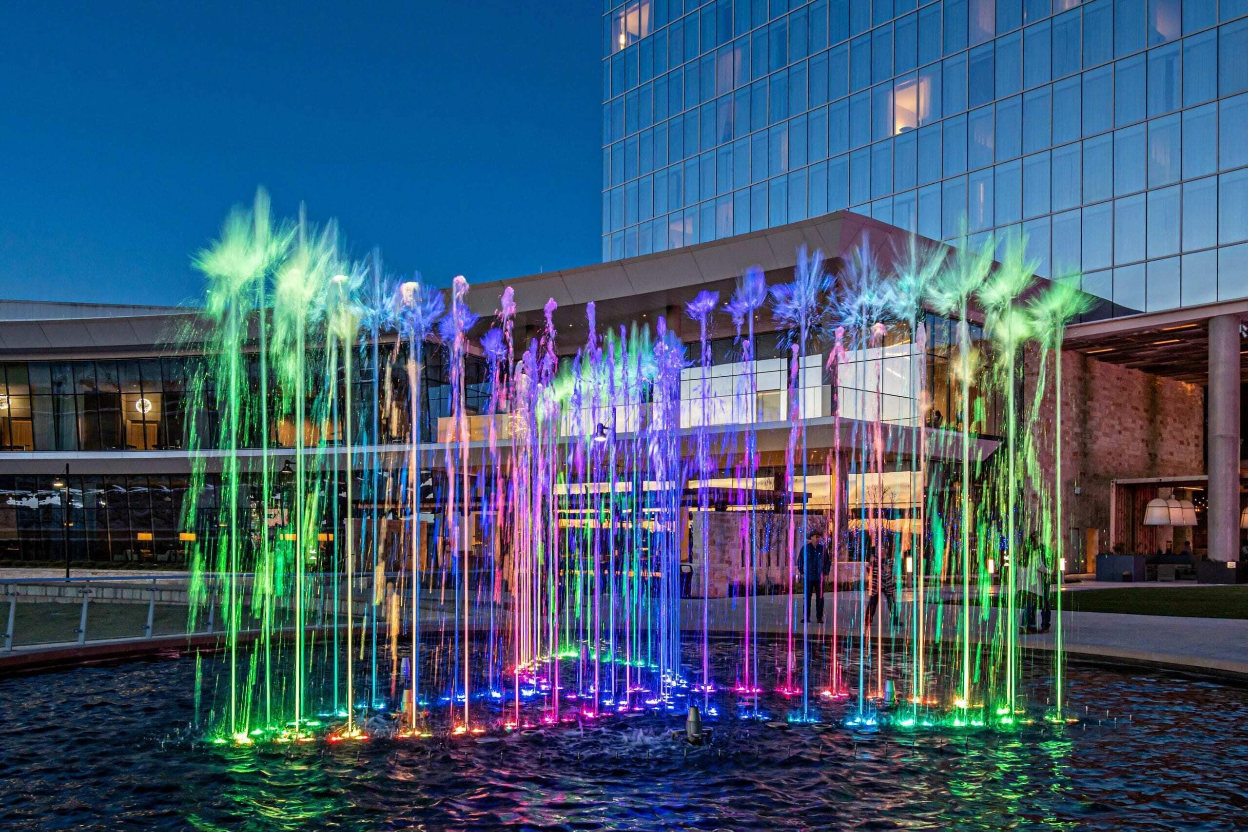 A nighttime view of a water fountain with colorful lights in front of a modern building.