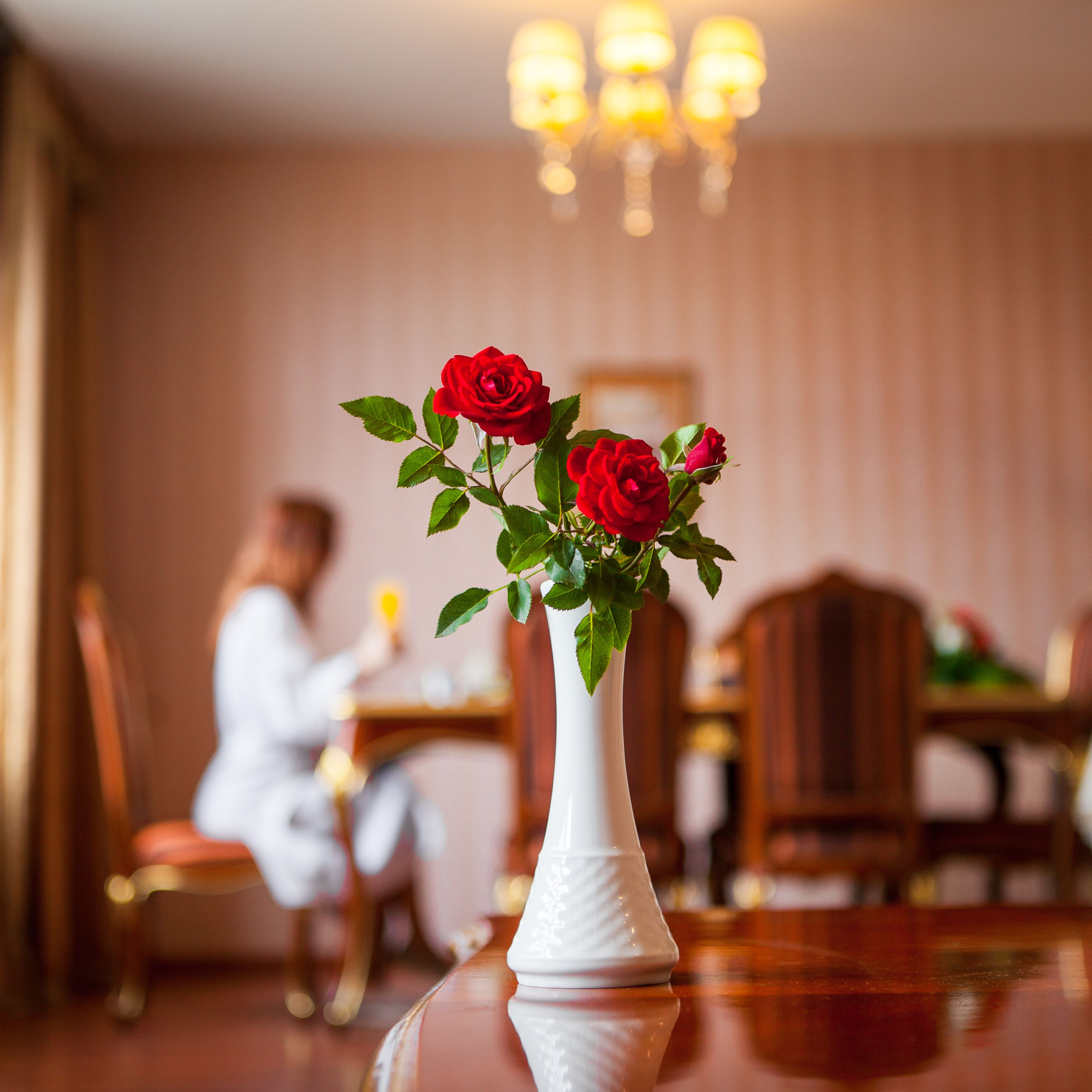 A white vase with red roses on a reflective surface in a dining area with wooden furniture and a chandelier in the background.