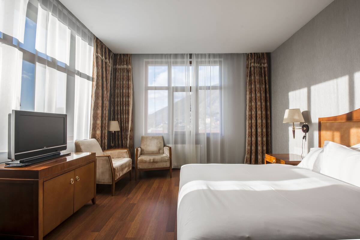 Une chambre d’hôtel avec un lit, un téléviseur posé sur une commode en bois, une chaise, un bureau et des rideaux.