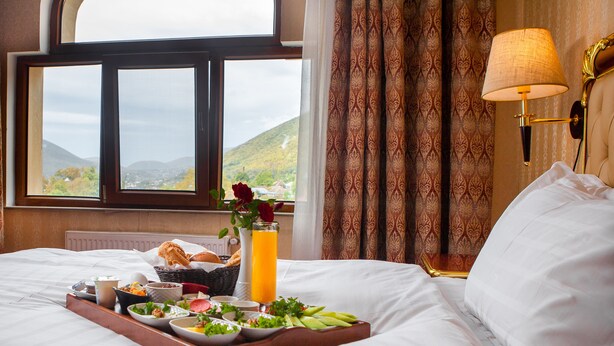 Una cama bien hecha con una bandeja de desayuno, un jarrón con una rosa roja y una vista de un paisaje montañoso a través de una ventana.