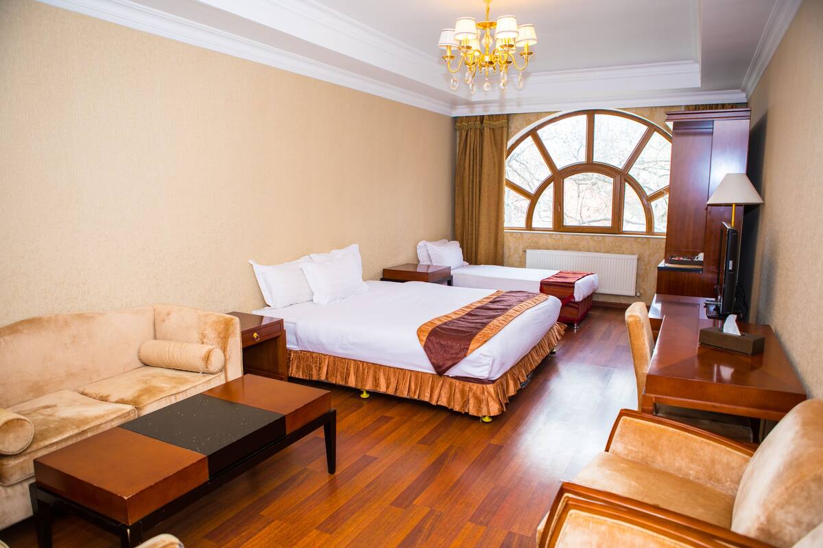 Une chambre d’hôtel avec un grand lit, un canapé, un bureau et une chaise. Il y a une fenêtre avec des rideaux et un lustre.