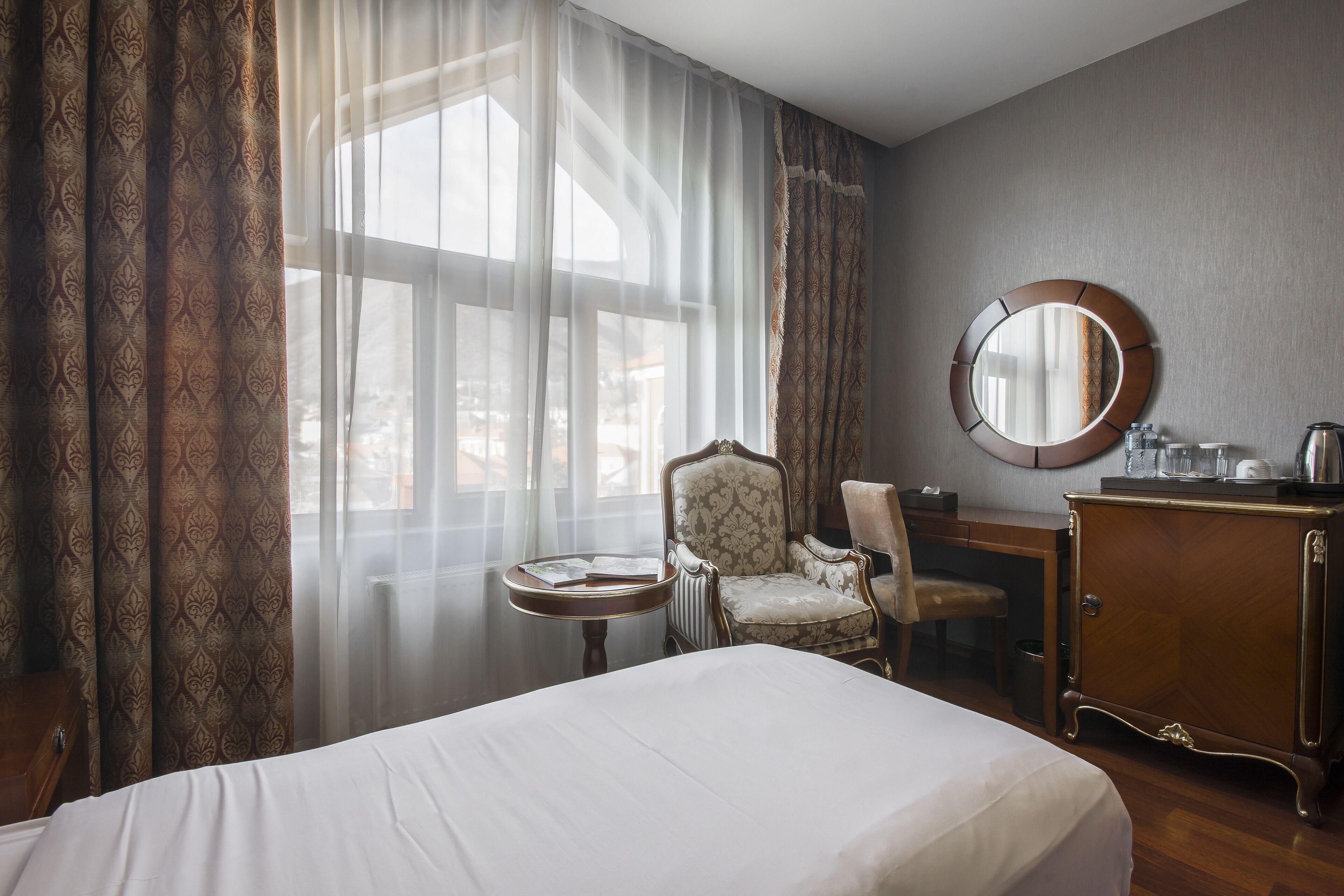 Habitación de hotel con una cama grande, un espejo redondo, un aparador de madera, una silla y una mesita.