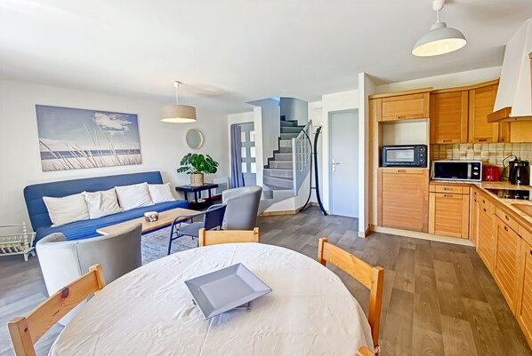 A modern living room with a blue sofa, a dining table with chairs, a staircase, and a kitchen area with wooden cabinets and appliances.