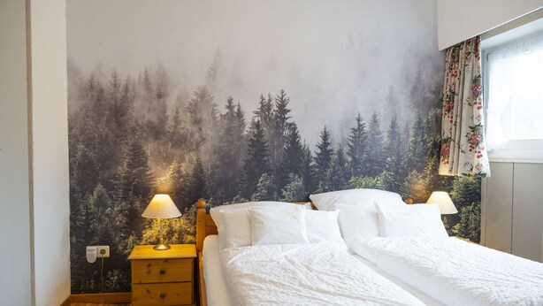 A bedroom with a forest-themed wall mural, a wooden bed with white bedding, two bedside lamps, and a small wooden nightstand with a lamp. - NATURION Hotel Hinterzarten (Hinterzarten)