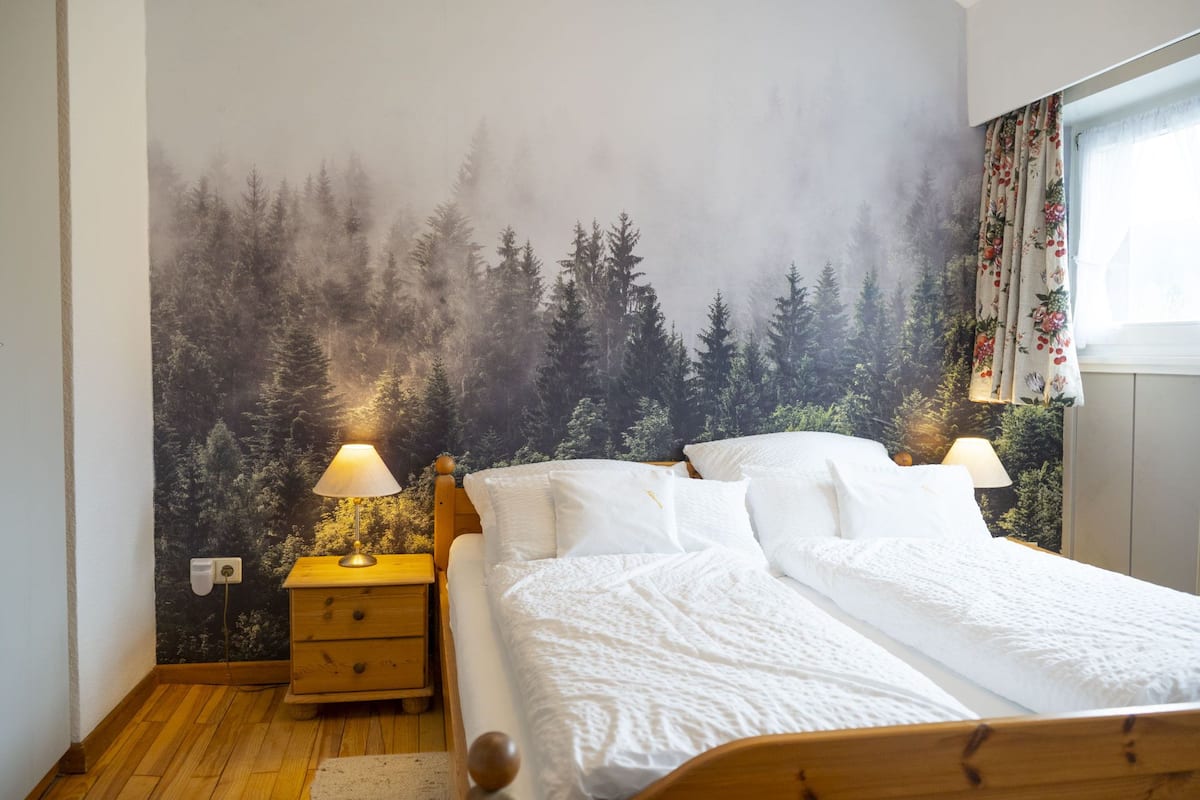 Ein Schlafzimmer mit Wandgemälde zum Thema Wald, einem hölzernen Bett mit weißer Bettwäsche, zwei Nachttischlampen und einem kleinen Nachttisch mit einer Lampe.