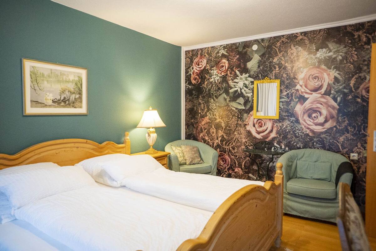 Ein Schlafzimmer mit Bett, Nachttisch, Lampe, Stuhl und floralem Wandpapier.