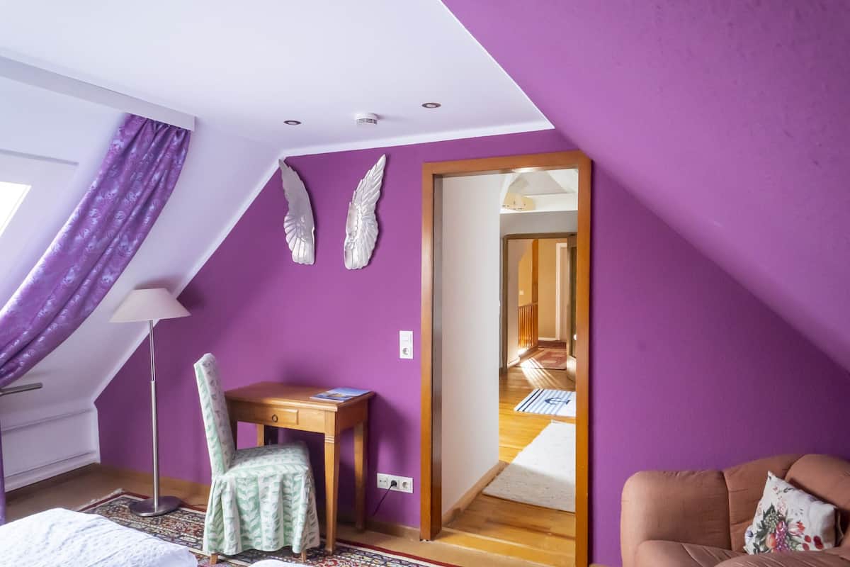 Ein Schlafzimmer mit schrägen Wänden, violetten Tapeten, einem Schreibtisch mit Stuhl, einem Bett und Blick durch eine offene Tür.