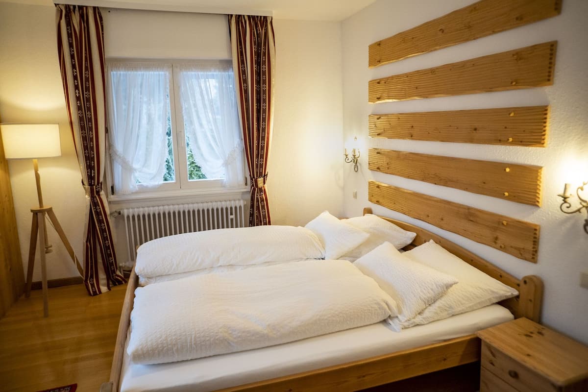 Ein Schlafzimmer mit einem hölzernen Kopfende, einem Bett mit weißen Leinen, einer Stehlampe und einem Fenster mit Vorhängen.