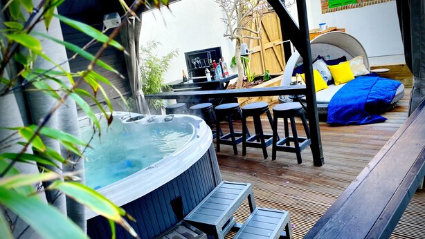 A hot tub with a wooden deck, a table with chairs, and a bed with pillows. - Ashbys (Portsmouth)