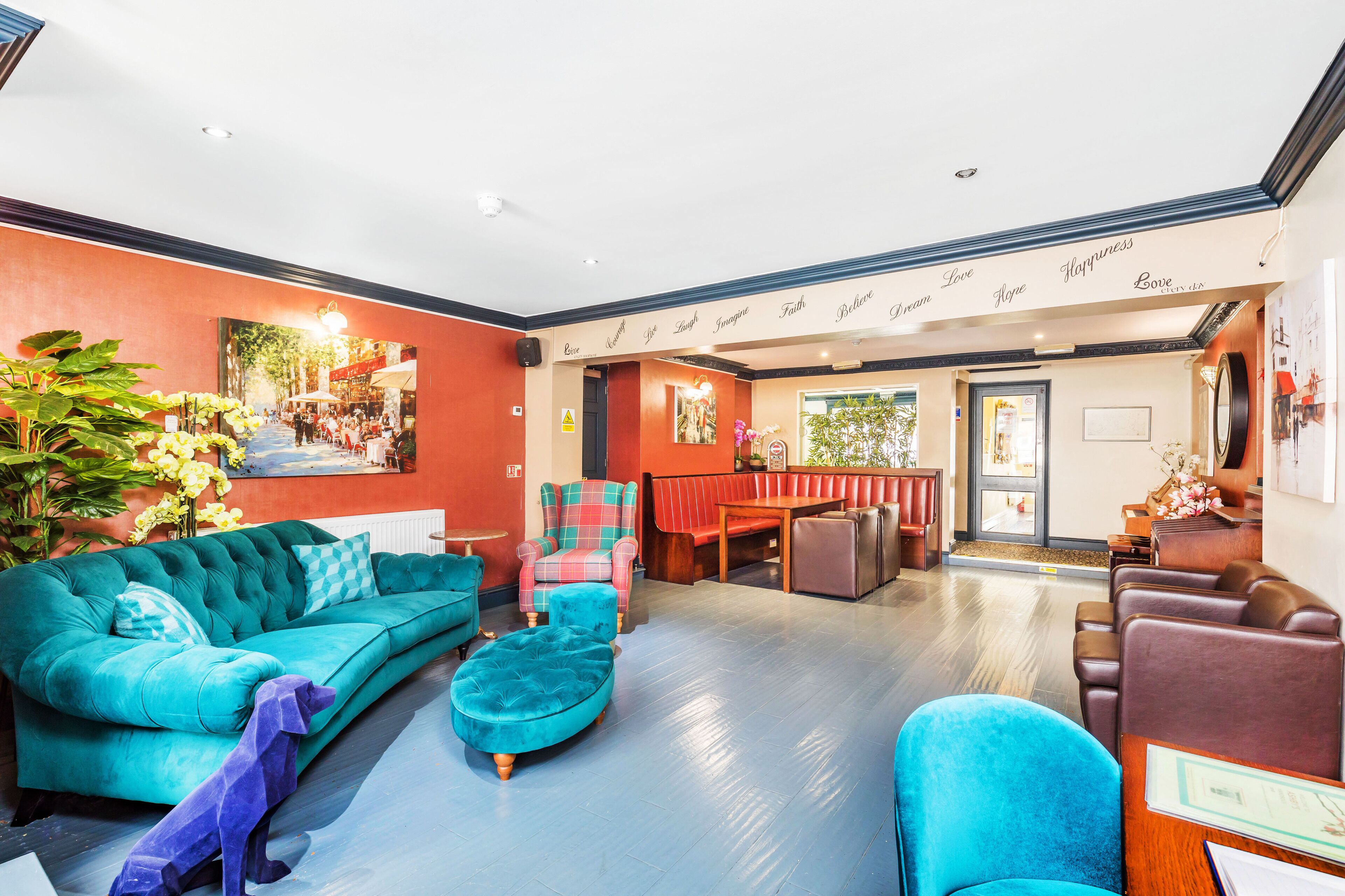 A hotel lobby with a teal sofa, a red and blue armchair, a wooden bench, and a reception area with a counter and a sign.