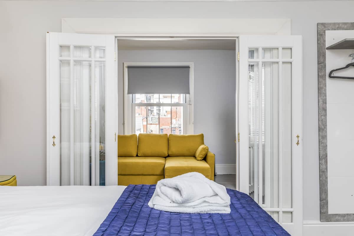 A hotel room with a bed, a yellow sofa, and a window with a view of buildings.