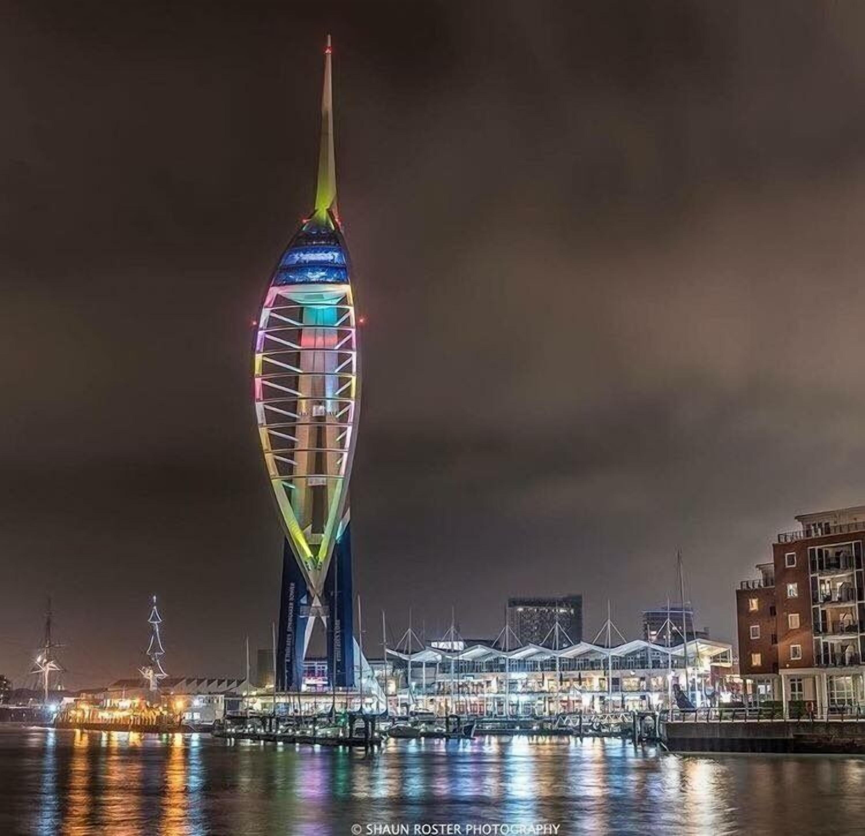 A tall, illuminated tower with a unique, curved design, surrounded by waterfront buildings and a marina.