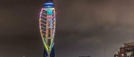 A tall, illuminated tower with a unique, curved design, surrounded by waterfront buildings and a marina.