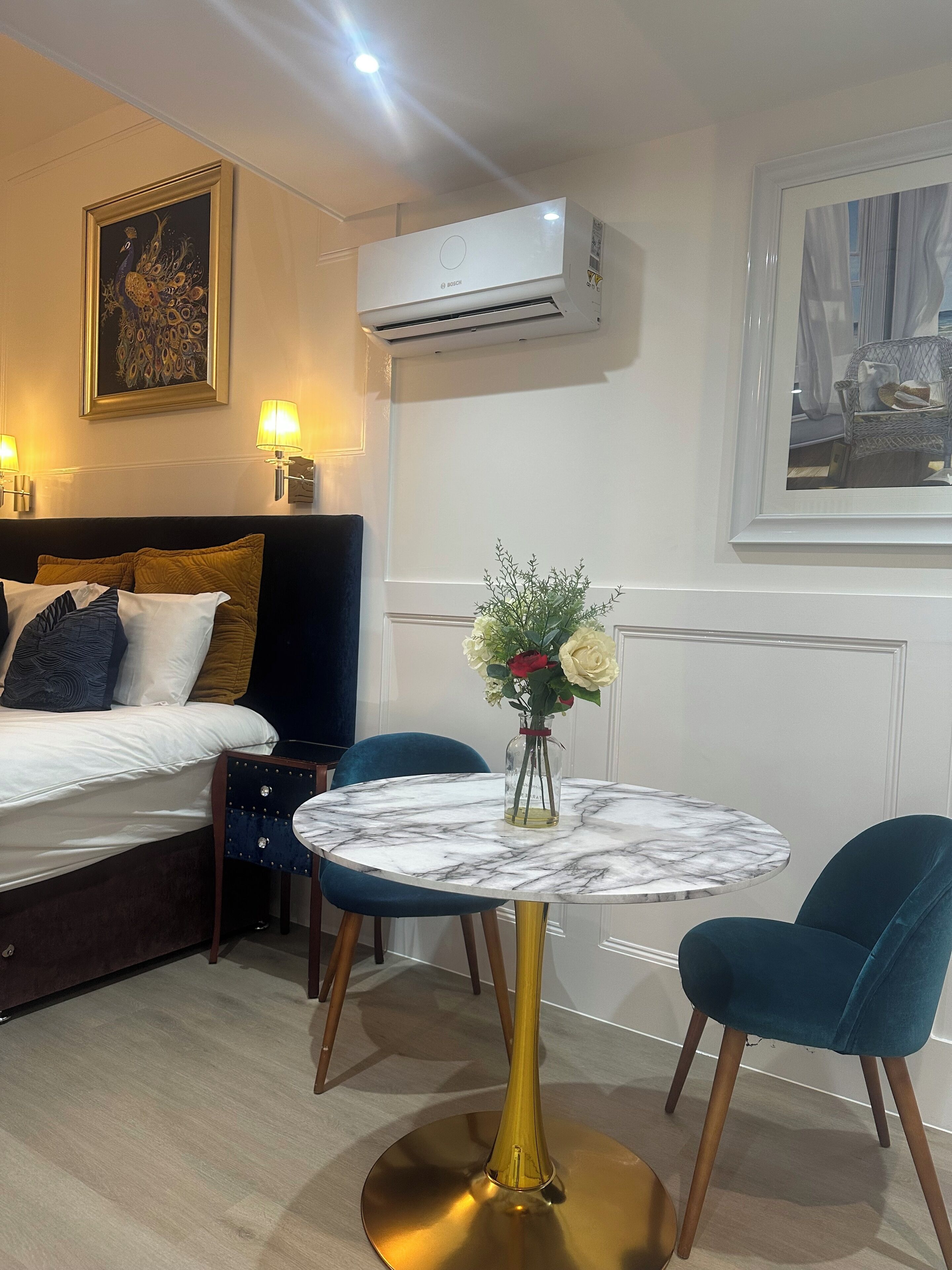 A hotel room with a bed, a round marble table with chairs, an air conditioning unit, and a framed artwork on the wall.