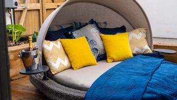 Un lit confortable surmonté d’un dais incurvé, une petite table avec une bouteille et un verre, et un fauteuil en osier.
