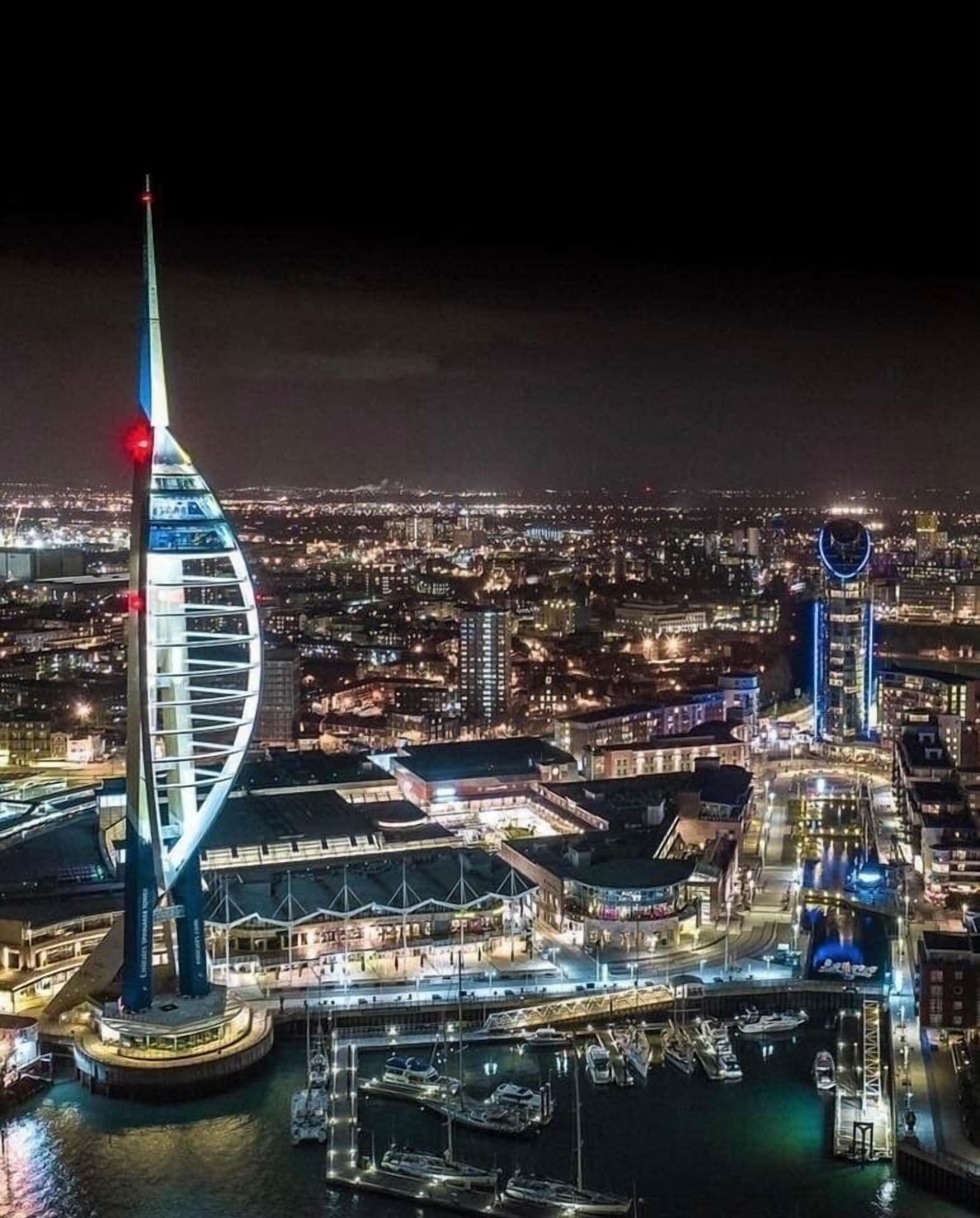 Une vue nocturne d’une ville, avec une tour imposante et illuminée, ainsi qu’un bâtiment circulaire doté d’un anneau bleu.