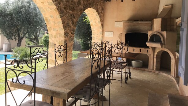 Un patio al aire libre con una mesa y sillas de madera, arcos de piedra, un horno de ladrillo y un colgador con luz.